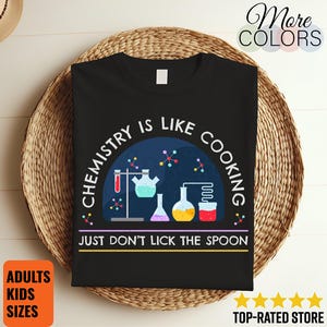 Könnte beinhalten: Schwarzes T-Shirt mit dem Text "CHEMISTRY IS LIKE COOKING JUST DON'T LICK THE SPOON" und wissenschaftlichen Grafiken. Das Design umfasst farbige Becher, Reagenzgläser und Molekülstrukturen. Das T-Shirt wird auf einem geflochtenen Korb präsentiert.