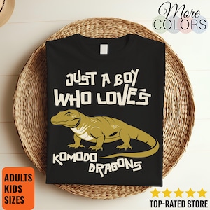 May include: Black t-shirt featuring the text "JUST A BOY WHO LOVES KOMODO DRAGONS" and a Komodo dragon graphic in green and yellow. The shirt is presented on a woven basket.