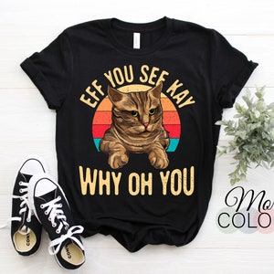 Könnte beinhalten: Schwarzes T-Shirt mit einer Katzen-Grafik in einem Retro-Sonnenuntergangskreis. Der Text lautet "EFF YOU SEE KAY WHY OH YOU". Schwarz-weiße Turnschuhe sind im Vordergrund. Rechts ist eine Pflanze.