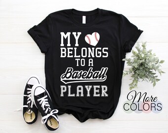 May include: Black t-shirt with the text "MY BELONGS TO A Baseball PLAYER" in white. A baseball heart graphic is above the text. A pair of black and white sneakers are in the foreground. A small plant is to the right.