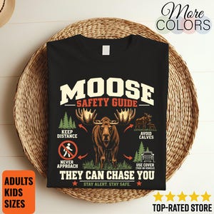 Może przedstawiać: Czarny t-shirt z grafiką zatytułowaną MOOSE SAFETY GUIDE. Projekt zawiera centralną ilustrację łosia otoczoną instrukcjami tekstowymi: KEEP DISTANCE, AVOID CALVES, NEVER APPROACH, USE COVER oraz THEY CAN CHASE YOU.