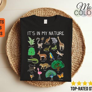 May include: Black t-shirt with the text "IT'S IN MY NATURE" and colorful illustrations of various animals and plants. The shirt includes a variety of animals such as a giraffe, koala, and alligator. The shirt is displayed on a woven basket.