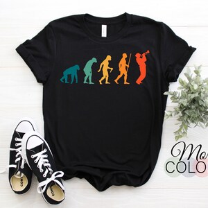 Puede incluir: Camiseta negra con un gráfico de la evolución del hombre, que termina con un hombre tocando la trompeta. El gráfico está en un esquema de colores retro. La camiseta está colocada sobre una superficie de madera blanca. Hay dos zapatillas negras y blancas en la esquina inferior izquierda de la imagen. El texto "Más colores" está en la esquina inferior derecha de la imagen.