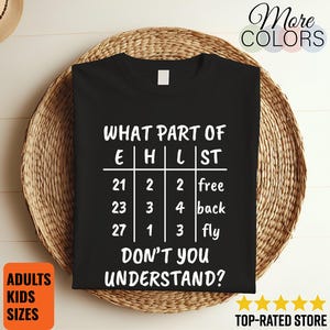 Puede incluir: Una camiseta negra de manga corta con texto blanco que dice: WHAT PART OF E H L ST 21 2 2 free 23 3 4 back 27 1 3 fly DON'T YOU UNDERSTAND? La camiseta se muestra sobre una alfombra redonda tejida, con un pequeño sombrero visible arriba a la izquierda.