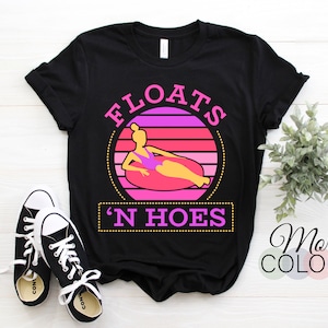 Może przedstawiać: Czarny T-shirt z różowo-żółtym nadrukiem przedstawiającym osobę na pontonie z tekstem "Floats 'N Hoes".