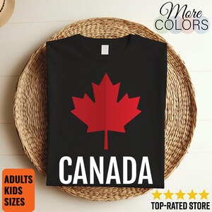 May include: A black t-shirt featuring a large red maple leaf graphic centered on the chest with the word CANADA printed in bold white capital letters underneath. The shirt has a simple crew neckline and short sleeves, presenting a clean design.