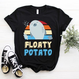 Puede incluir: Camiseta negra con un diseño de foca de dibujos animados y el texto "FLOATY POTATO". La foca es de color azul claro, sobre un fondo de puesta de sol en rojo, naranja y amarillo. Zapatillas deportivas blancas y negras en primer plano.
