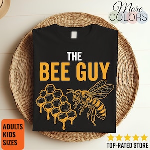 May include: A black t-shirt featuring the white text THE and the yellow text BEE GUY above a yellow graphic illustration of a honeycomb with dripping honey and a honeybee. The design is centered on the front of the shirt.
