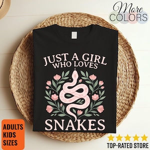 Puede incluir: Una camiseta negra con un estampado gráfico de una serpiente rosa claro enroscada entre flores rosa claro y hojas verde salvia. El texto "JUST A GIRL WHO LOVES SNAKES" está impreso en letras mayúsculas de color rosa claro sobre la ilustración.