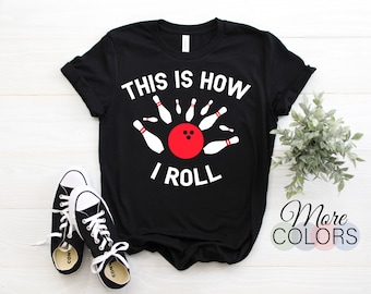 May include: Black t-shirt with the text "THIS IS HOW I ROLL" in white. The design features a red bowling ball surrounded by white bowling pins. A pair of black and white sneakers are in the foreground. The shirt is displayed on a white wooden surface.