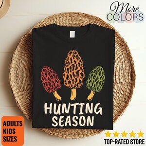 May include: Black t-shirt featuring three colorful morel mushrooms in red, orange, and green. The words "HUNTING SEASON" are printed in white below the mushrooms. The shirt is displayed on a woven basket.