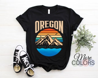 Könnte beinhalten: Schwarzes T-Shirt mit einem kreisförmigen Grafikdesign, das das Wort "OREGON" in einem beigen Farbton, eine Bergkette, einen Sonnenuntergang und den Text "PACIFIC NORTHWEST" zeigt. Schwarze und weiße Turnschuhe sind im Vordergrund.