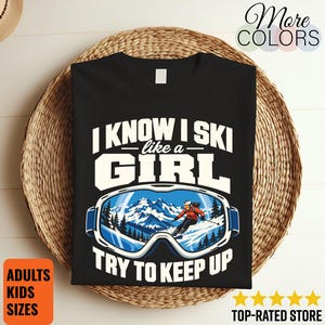 May include: A black t-shirt featuring white text that reads I KNOW I SKI like a GIRL TRY TO KEEP UP. The graphic includes a pair of ski goggles with a detailed illustration of a person skiing down a snowy mountain landscape inside the lenses.
