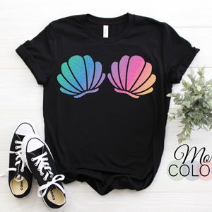 Può includere: Maglietta nera con un design di due conchiglie arcobaleno con glitter. Il testo "More Colors" è visibile sotto la maglietta.