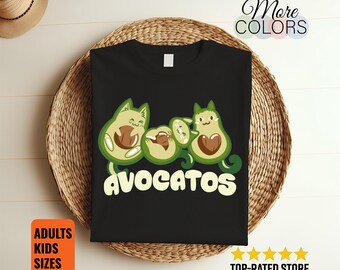 May include: Black t-shirt featuring a whimsical design of avocado cats with the word "AVOCATOS" in white. The avocados have cat-like features and are in shades of green and brown. The shirt is displayed on a woven surface.