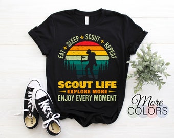 May include: Black t-shirt with a graphic design featuring a silhouette of a hiker against a sunset backdrop. The text reads "EAT SLEEP SCOUT REPEAT" and "SCOUT LIFE EXPLORE MORE ENJOY EVERY MOMENT". Black and white sneakers are in the foreground.