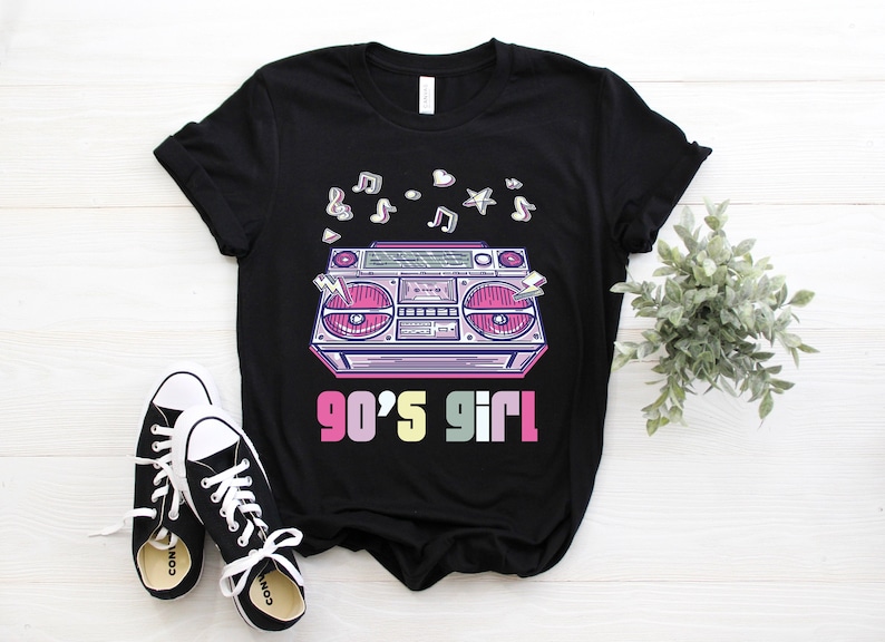 May include: Black t-shirt featuring a retro boombox graphic with musical notes and the text "90's Girl". The design includes pink, purple, and yellow accents. A pair of black and white sneakers are in the foreground.