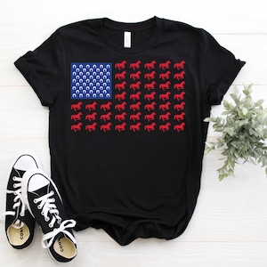 Könnte beinhalten: Schwarzes T-Shirt mit einem rot-weiß-blauen amerikanischen Flaggen-Design. Die Flagge besteht aus roten Hufeisen und roten Pferden, die in Richtung der Flaggenstange laufen.
