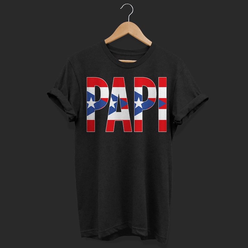 May include: Black t-shirt with the word "PAPI" in large, bold letters. The letters are designed with the Puerto Rican flag's colors: red, white, and blue, with stars. The shirt is displayed on a wooden hanger.