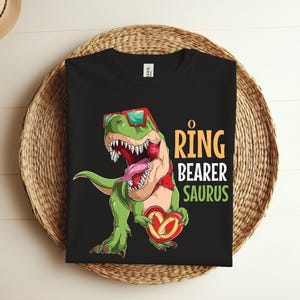 May include: Black t-shirt with a green dinosaur wearing sunglasses and holding a heart-shaped ring. The text "O Ring Bearer Saurus" is printed on the shirt.