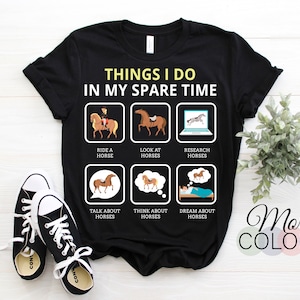 Op de afbeelding: Zwart T-shirt met een witte afbeelding waarop staat "Things I do in my spare time" en een reeks illustraties van een persoon die paardrijdt, naar paarden kijkt, onderzoek doet naar paarden, over paarden praat, aan paarden denkt en van paarden droomt.