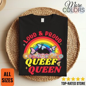 Puede incluir: Camiseta negra con el texto "LOUD & PROUD QUEEF QUEEN" en rojo y amarillo. El diseño incluye un arcoíris, mariposas y flores. La camiseta se muestra sobre una cesta de mimbre.