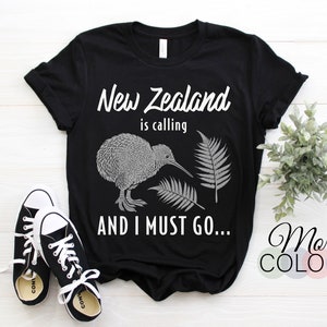 Może przedstawiać: Czarny T-shirt z białym nadrukiem kiwi i liści paproci. Na koszulce widnieje napis "New Zealand is calling and I must go..."
