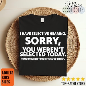 Könnte beinhalten: Schwarzes T-Shirt mit dem Text "I HAVE SELECTIVE HEARING. SORRY, YOU WEREN'T SELECTED TODAY. TOMORROW ISN'T LOOKING GOOD EITHER." Das Shirt wird auf einem geflochtenen Korb präsentiert. Zusätzlicher Text enthält "ADULTS KIDS SIZES" und "TOP-RATED STORE."