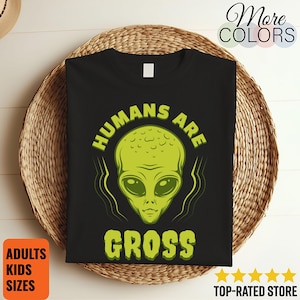 May include: Black t-shirt with a green alien graphic and the text "HUMANS ARE GROSS". The alien has large eyes and a bulbous head. The shirt is displayed on a woven surface. The shirt is available in adult and kids sizes.