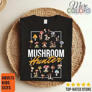 May include: Black t-shirt featuring a collection of colorful mushroom illustrations within a white rectangular frame. The words "MUSHROOM Hunter" are printed in white and gold. The shirt is displayed on a woven basket.