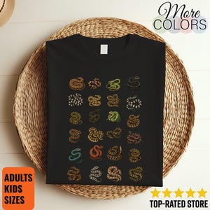 Puede incluir: Una camiseta negra con una cuadrícula de veinticuatro pequeñas ilustraciones detalladas de serpientes en varios colores y patrones, incluyendo verde, naranja, marrón y negro, dispuestas en seis filas de cuatro en el centro.