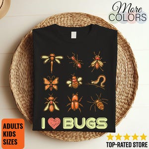 Peut inclure: T-shirt noir avec des illustrations colorées de divers insectes, dont une libellule, une fourmi, un papillon, une abeille et une araignée. Le texte "I ❤️ BUGS" est imprimé en dessous. Le t-shirt est présenté sur un panier tressé.