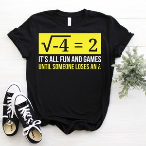 May include: Black t-shirt with a yellow rectangle containing the equation "√-4 = 2". Below the equation is the text "IT'S ALL FUN AND GAMES UNTIL SOMEONE LOSES AN i.". Black and white sneakers are at the bottom.