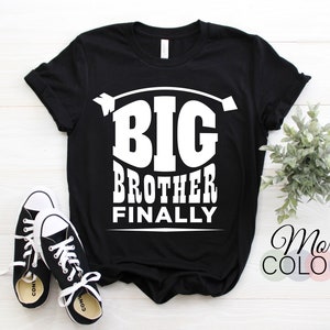 Puede incluir: Camiseta negra con el texto blanco "BIG BROTHER FINALLY". La camiseta se combina con zapatillas deportivas en blanco y negro. Una pequeña planta y una paleta de colores están en el fondo.