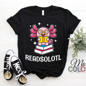 May include: Black t-shirt featuring a cartoon axolotl wearing glasses and reading a book, with the text "READSOLOTL". The axolotl is pink with a white outline and is sitting on top of a stack of books. Black and white sneakers are in the foreground.