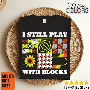 May include: Black t-shirt with the text "I STILL PLAY WITH BLOCKS" in white. The shirt features a geometric design in orange, green, and white. The shirt is displayed on a woven surface. The words "ADULTS KIDS SIZES" are in a square orange box.