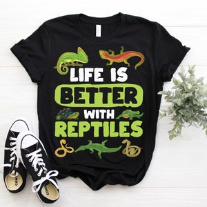 Op de afbeelding: Zwart T-shirt met de tekst "LIFE IS BETTER WITH REPTILES" en reptielenillustraties. Het ontwerp bevat een groene kameleon, een oranje hagedis, een schildpad, een krokodil en een slang. Zwarte en witte sneakers staan op de voorgrond.