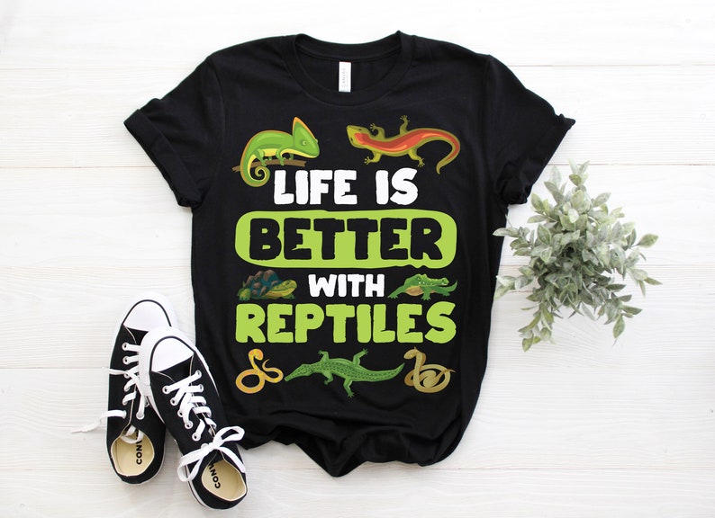以下が含まれることがあります： 「LIFE IS BETTER WITH REPTILES」の文字と爬虫類のイラストが描かれた黒いTシャツ。デザインには、緑のカメレオン、オレンジ色のトカゲ、カメ、ワニ、ヘビが含まれています。黒と白のスニーカーが前景にあります。