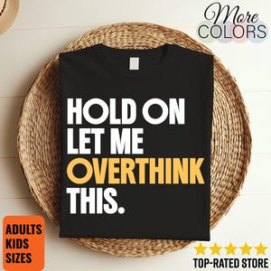 May include: A black short-sleeved t-shirt featuring the white text "HOLD ON LET ME" and the yellow text "OVERTHINK" followed by the white text "THIS." The shirt has a simple crew neckline and is displayed flat, showcasing the bold graphic design.