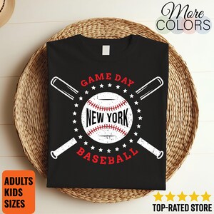 Op de afbeelding: Zwart T-shirt met een honkbalontwerp. Het ontwerp bevat een honkbal met "NEW YORK" in het midden, omringd door sterren en gekruiste honkbalknuppels. De woorden "GAME DAY" en "BASEBALL" staan ook in het ontwerp. Het shirt ligt op een geweven oppervlak.