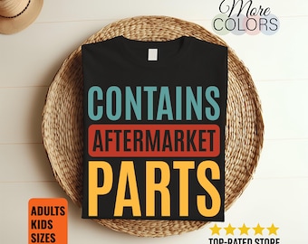 May include: Black t-shirt with the text "CONTAINS AFTERMARKET PARTS" in teal, red, and yellow. The shirt is displayed on a woven basket. Additional text includes "More Colors", "Adults Kids Sizes", and "Top-Rated Store".