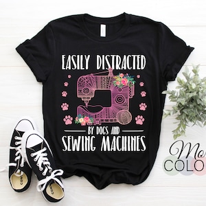 Peut inclure: T-shirt noir avec l'inscription "EASILY DISTRACTED by DOGS and SEWING MACHINES". Le motif comprend une machine à coudre rose, des empreintes de pattes, des fleurs et une pelote de laine. Une paire de baskets noires et blanches est au premier plan.