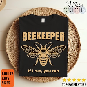 May include: Black t-shirt with a bee graphic and the word "BEEKEEPER" in a distressed font. Below the bee illustration, the text reads "If I run, you run." The shirt is displayed on a woven basket.