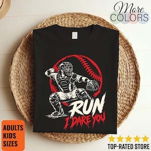 May include: A black t-shirt featuring a graphic of a baseball catcher in a crouched position in front of a red baseball silhouette. The text RUN I DARE YOU is printed in white and red below the catcher. The design is centered on the shirt.