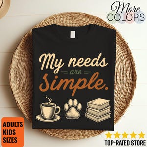 Puede incluir: Una camiseta negra con el texto My needs are Simple en letras color crema y marrón. Debajo del texto hay tres ilustraciones: una taza de café humeante sobre un plato, una huella de pata y una pila de tres libros, todo en color crema.