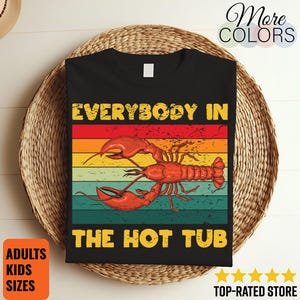 May include: Black t-shirt with a retro design featuring a red lobster and the text "EVERYBODY IN THE HOT TUB" in yellow. The design has a vintage sunset colour scheme. The shirt is displayed on a woven basket.