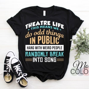 Op de afbeelding: Zwart T-shirt met witte tekst met de tekst "THEATRE LIFE THIS MEANS WE do odd things IN PUBLIC HANG WITH WEIRD PEOPLE RANDOMLY BREAK INTO SONG." Het shirt wordt gecombineerd met zwart-witte sneakers. De woorden "More COLORS" zijn ook zichtbaar.