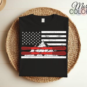 Op de afbeelding: Zwart T-shirt met een verweerd Amerikaans vlagontwerp met een bergketen en twee rode ski's. De tekst "More Colors" is zichtbaar in de rechterbovenhoek van de afbeelding.