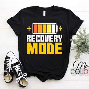 May include: Black t-shirt featuring the text "RECOVERY MODE" in white and yellow, with a battery graphic. The image includes black and white trainers. A small plant and the words "More Colors" are also visible.