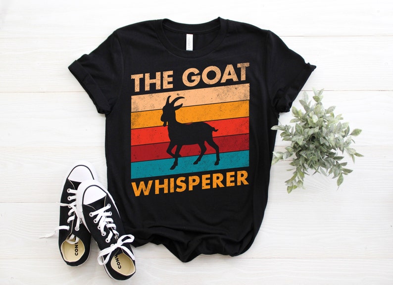 Puede incluir: Camiseta negra con el texto "THE GOAT WHISPERER" encima y debajo de una silueta de cabra. El texto y la cabra est&aacute;n sobre un fondo retro de rayas naranjas, amarillas, rojas y turquesas. Zapatillas de lona negras con cordones blancos en primer plano.